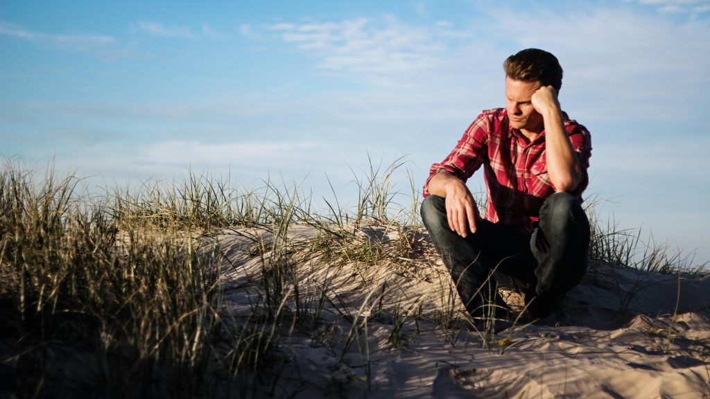 Homme en chemise à carreaux assis pensivement sur une dune de sable avec de l'herbe